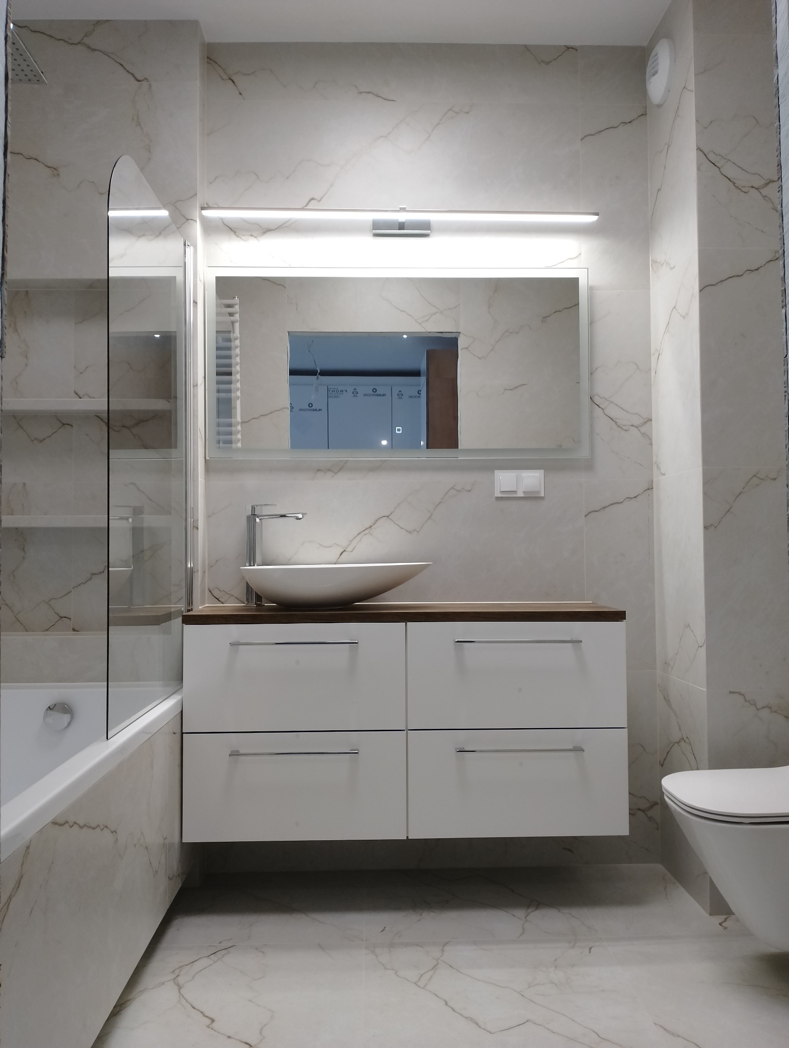 Nowoczesna łazienka z marmurowymi płytkami, białą szafką pod umywalką i lustrem z oświetleniem LED. Widoczna wanna z parawanem i toaleta w minimalistycznym stylu.