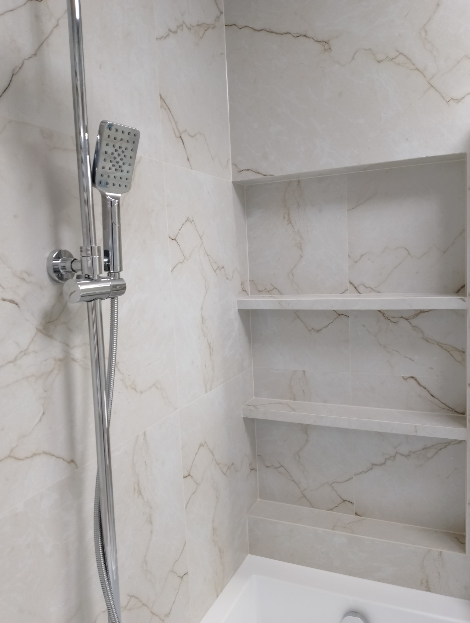 Wykończona kabina prysznicowa z marmurowymi płytkami i wnękami na kosmetyki. Chromowana bateria natryskowa. Minimalistyczny design, jasne wnętrze.