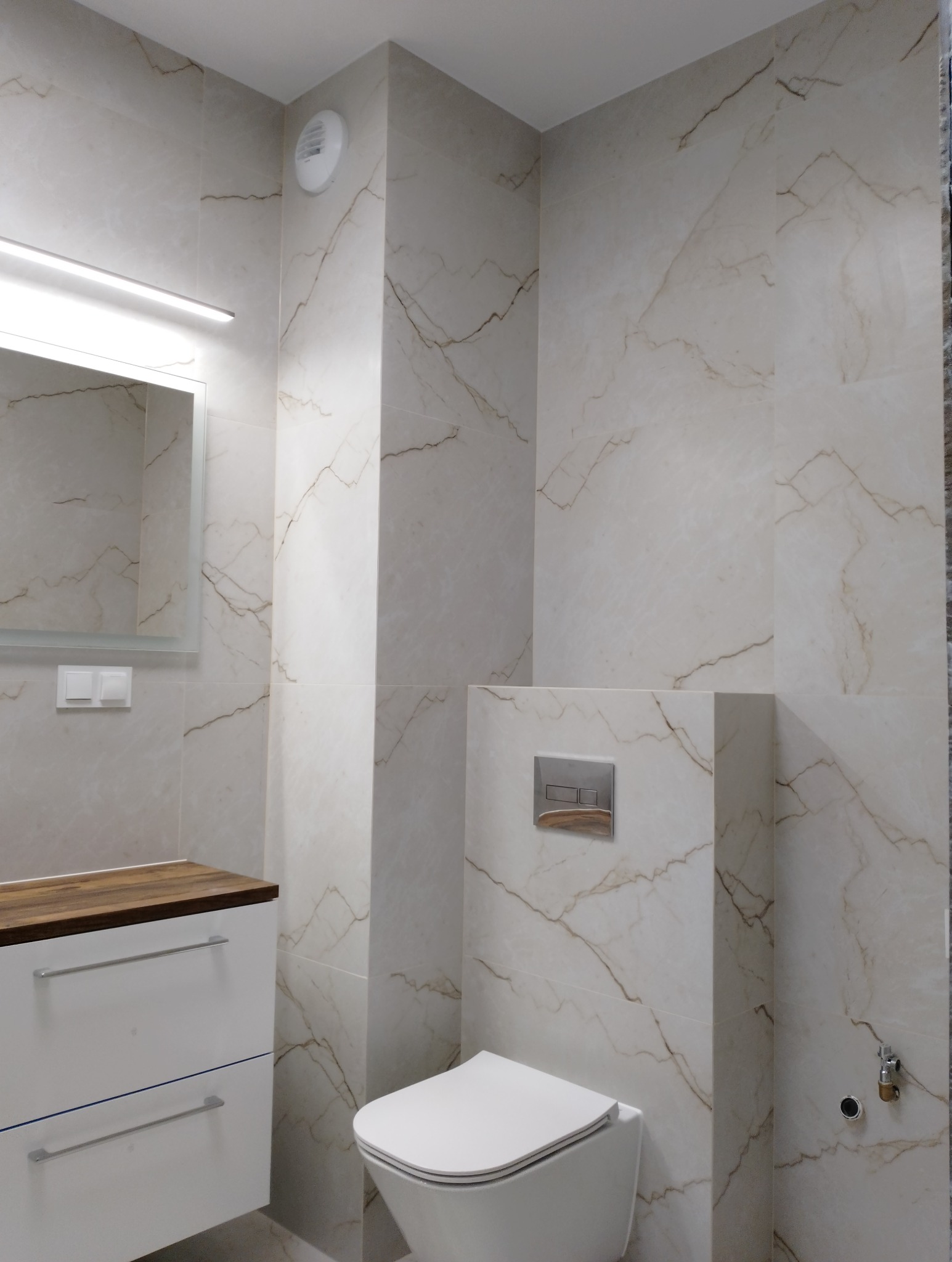 Nowoczesna łazienka z marmurowymi płytkami, podwieszaną toaletą i szafką z drewnianym blatem. Minimalistyczny design, oświetlenie LED nad lustrem, wentylator sufitowy.