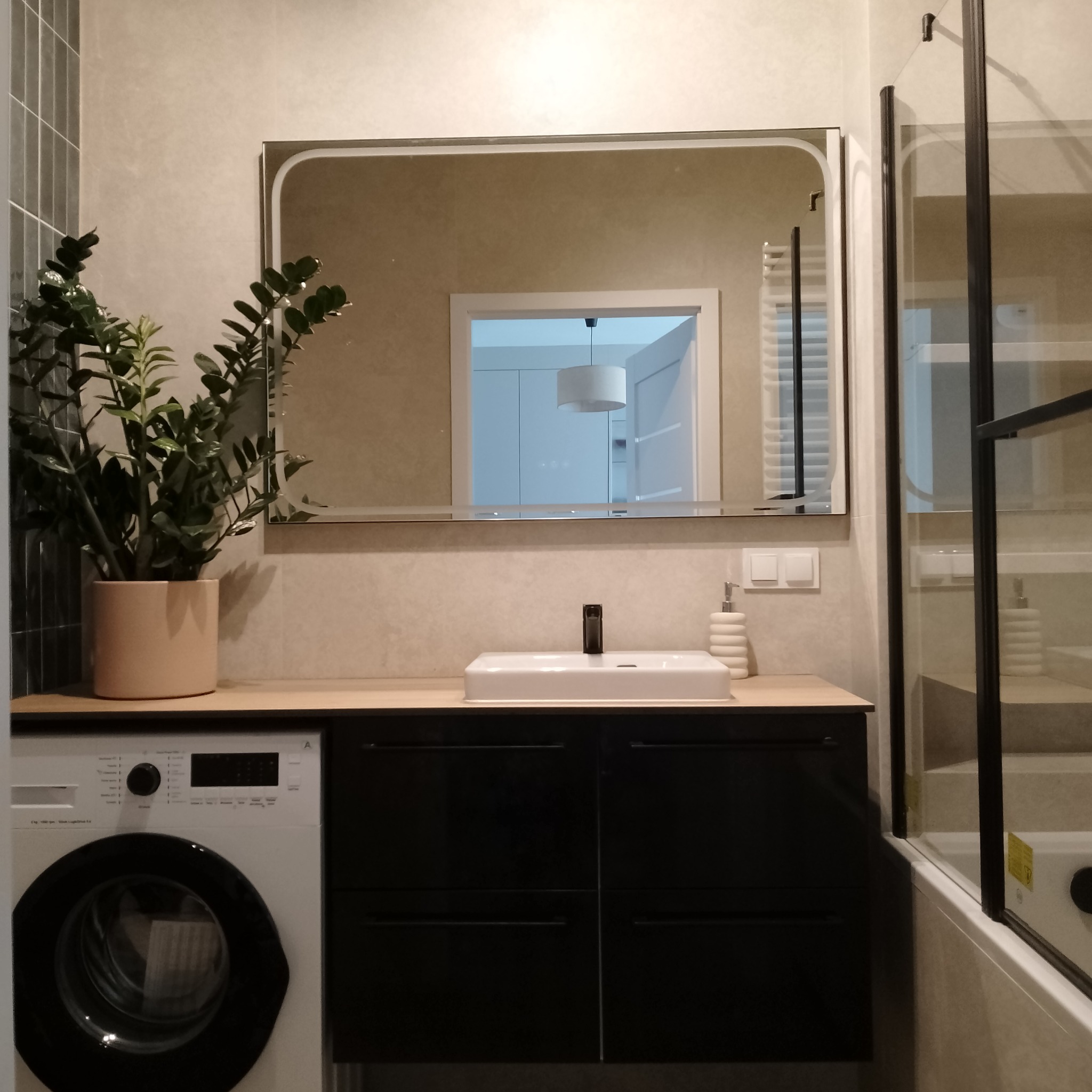 Jasna łazienka z umywalką na szafce, pralką i rośliną w doniczce. W lustrze odbija się widok na inne pomieszczenie. Kabina prysznicowa z czarnymi ramami.