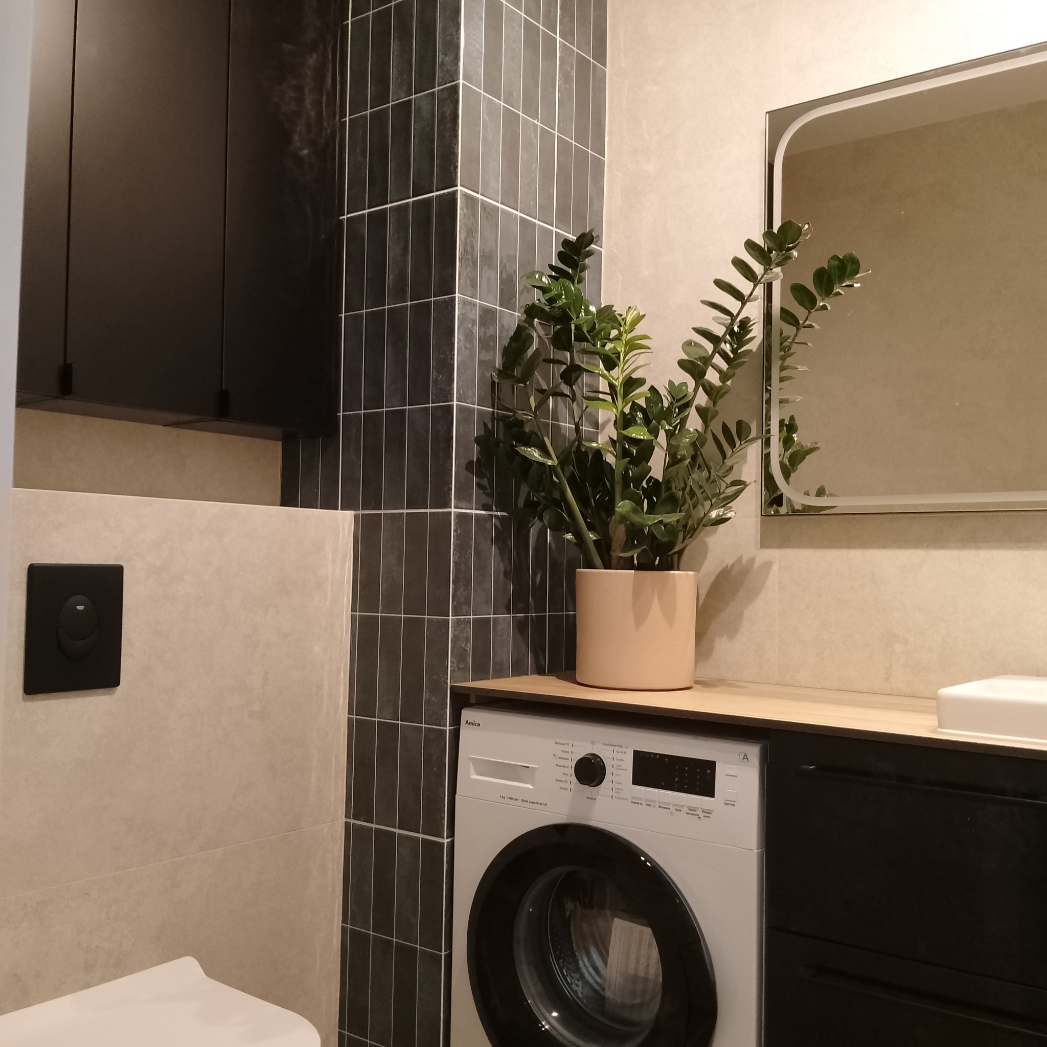 Nowoczesna łazienka z ciemnymi, pionowo ułożonymi płytkami, pralką Amica i rośliną w doniczce. Minimalistyczny design z elementami drewna i czerni.