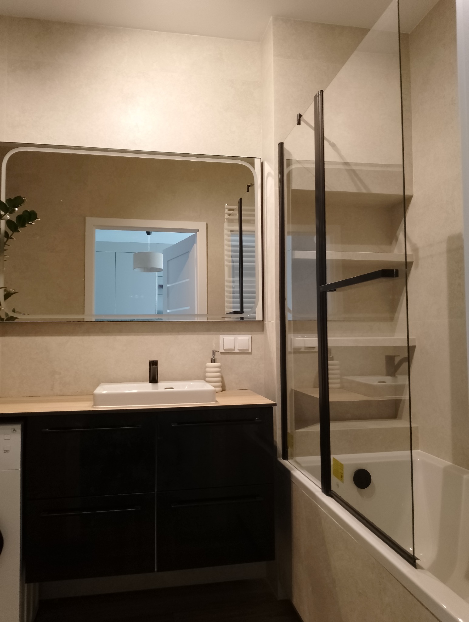 Nowoczesna łazienka po remoncie: czarna szafka z drewnianym blatem, umywalka, lustro z zaokrąglonymi rogami i szklany parawan nawannowy z czarnymi ramami.