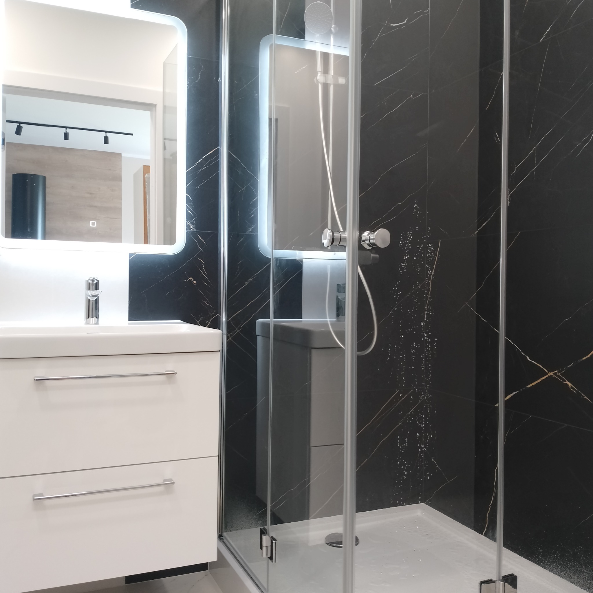 Nowoczesna łazienka z czarnymi płytkami imitującymi marmur, białą szafką z umywalką i podświetlanym lustrem oraz kabiną prysznicową ze szklanymi drzwiami.