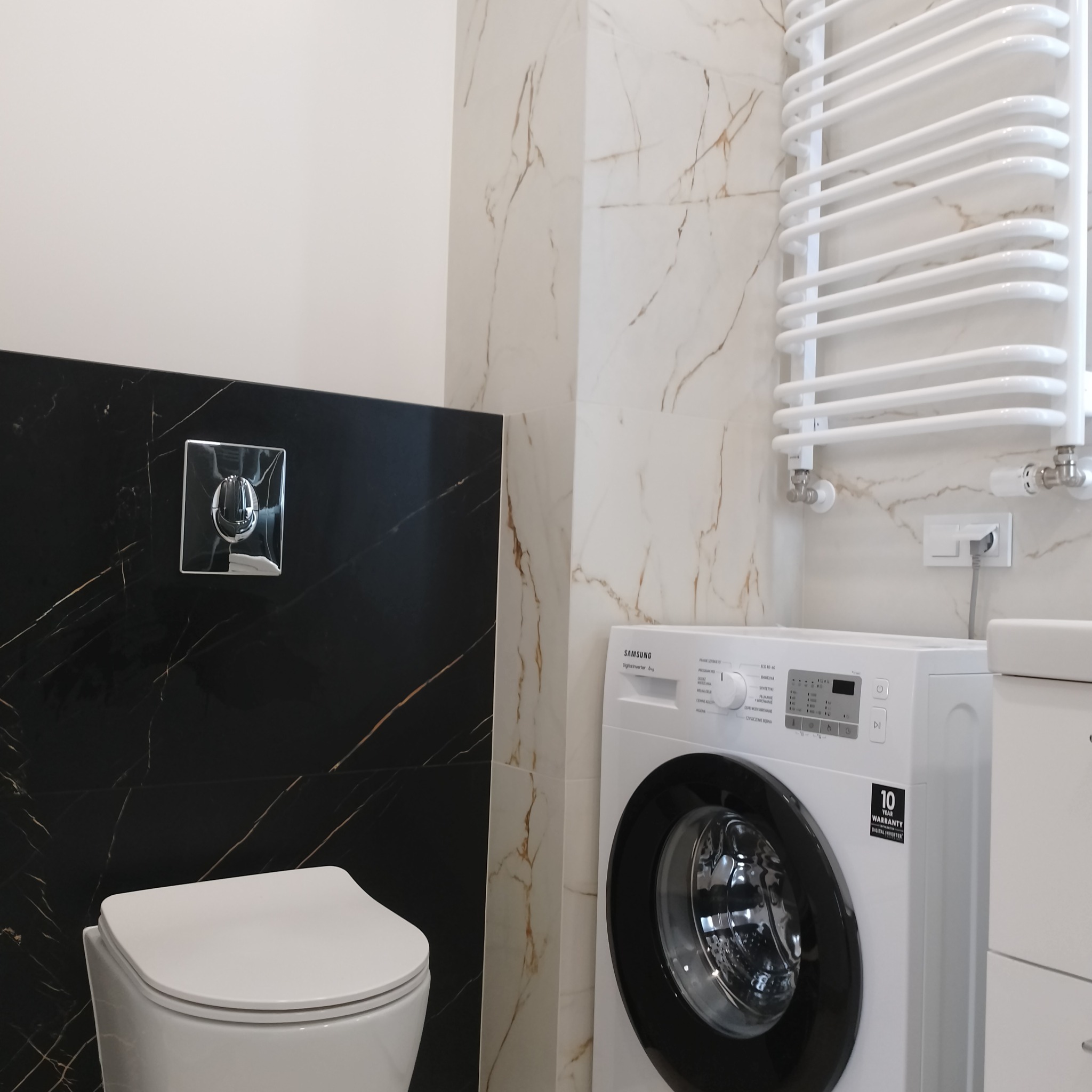 Nowoczesna łazienka z czarnymi i białymi płytkami imitującymi marmur, wisząca toaleta, grzejnik drabinkowy i pralka Samsung. Minimalistyczny design.
