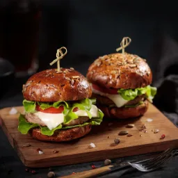 Dwa apetyczne burgery na drewnianej desce, jeden z posypką z ziaren, drugi z widocznymi nasionami na bułce, oba spięte ozdobnymi wykałaczkami, z widocznymi warstwami sałaty, pomidora i sera,...