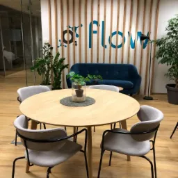 Jasne wnętrze biurowe z okrągłym stołem, czterema szarymi krzesłami i niebieską sofą na tle ściany z logo 'Flow' i drewnianymi panelami.