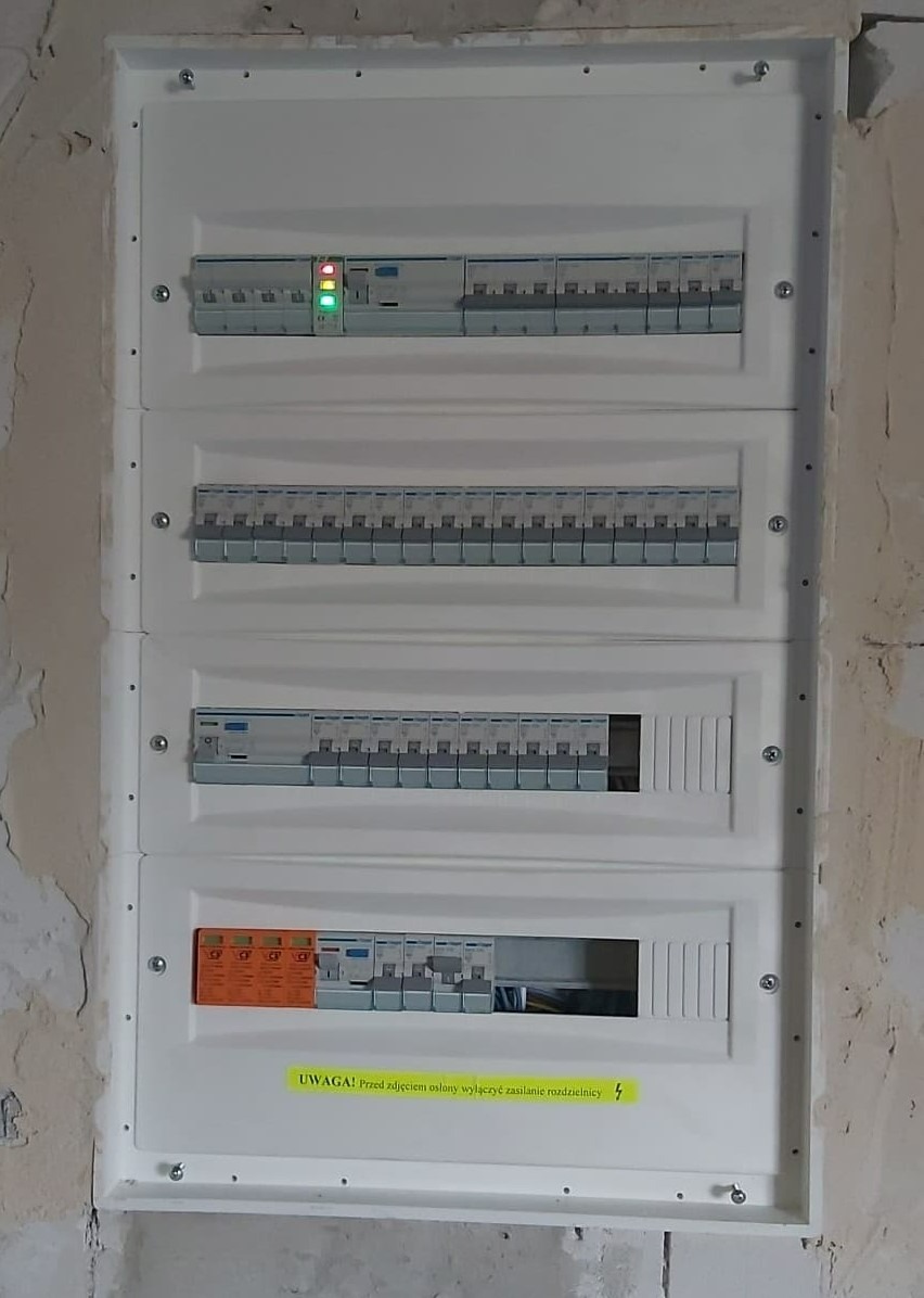 Rozdzielnica elektryczna zamontowana na surowej ścianie w Leńczach, widoczne bezpieczniki i wyłączniki różnicowoprądowe, z naklejką ostrzegawczą.