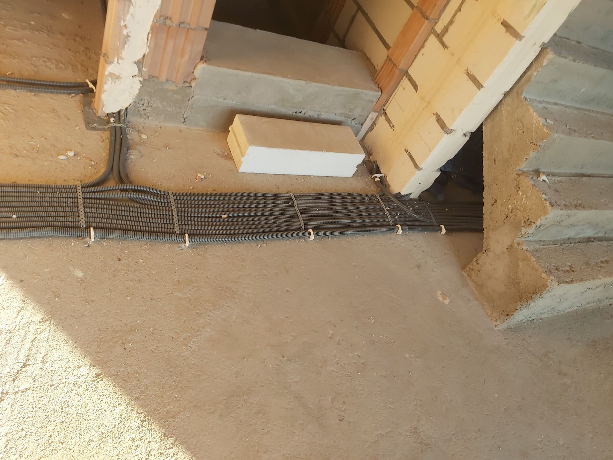 Rzędy szarych rur karbowanych, przymocowanych białymi uchwytami do surowego betonowego podłoża w trakcie budowy, biegnące wzdłuż schodów z betonu i cegieł.