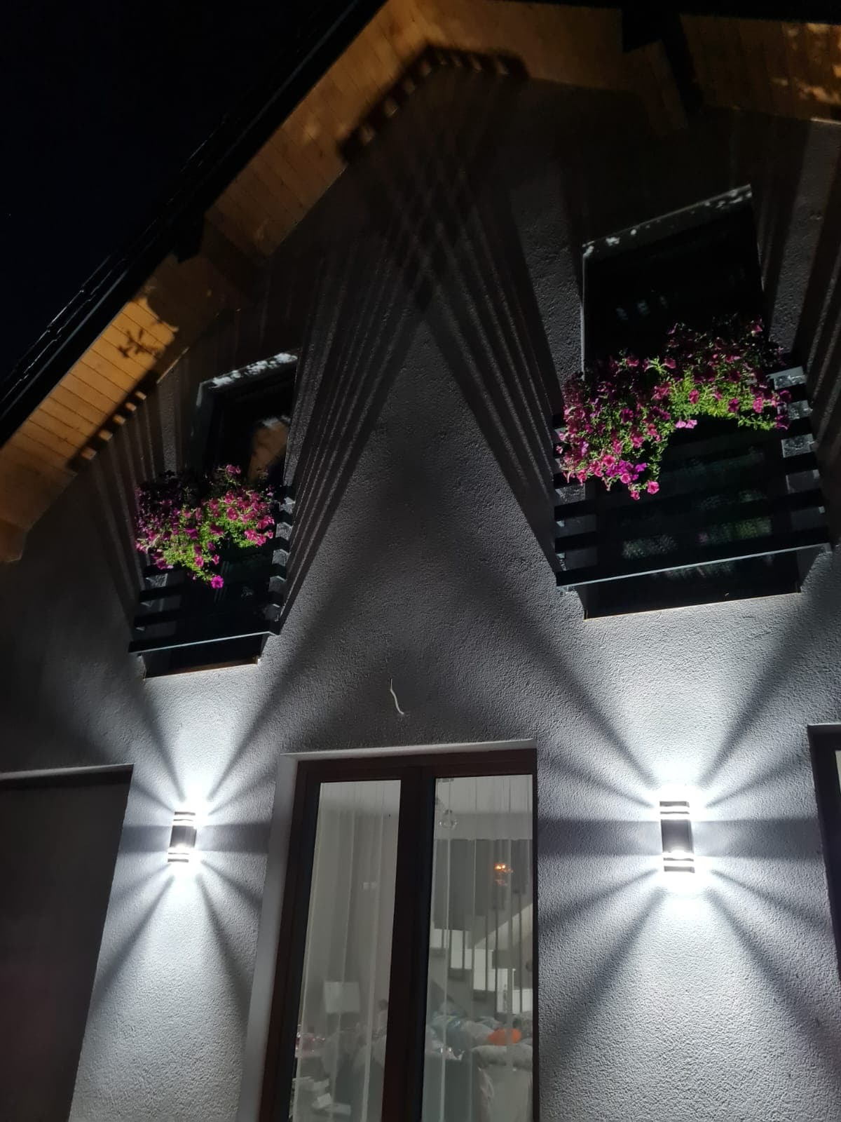 Elewacja domu oświetlona dwoma nowoczesnymi kinkietami, z drewnianymi skrzynkami kwiatowymi pod oknami mansardowymi w nocnym ujęciu.