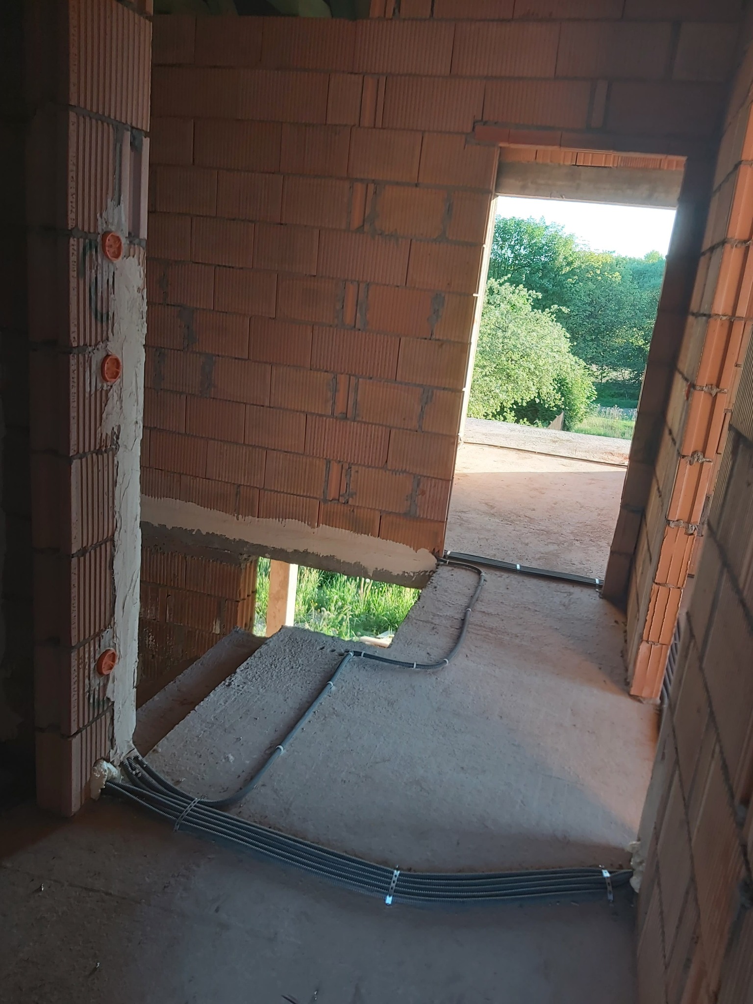 Wnętrze w trakcie budowy z widocznymi przewodami elektrycznymi w peszlach, prowadzonymi wzdłuż ściany z cegły ceramicznej, z puszkami instalacyjnymi wmurowanymi w ścianę i widokiem na zieleń przez...