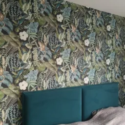 Ściana sypialni pokryta tapetą z motywem dżungli z małpami, kwiatami i liśćmi w odcieniach zieleni, niebieskiego i brązu, widoczny fragment wezgłowia łóżka w kolorze butelkowej zieleni i fragment...