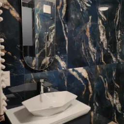 Nowoczesna łazienka z umywalką nablatową o geometrycznym kształcie, bateria chromowana, ściany wyłożone płytkami imitującymi marmur w odcieniach granatu i złota, owalne lustro z podświetleniem LED.