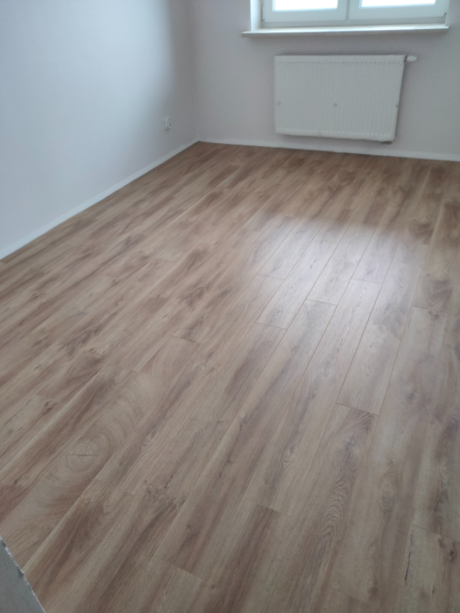 Jasnobrązowe panele podłogowe ułożone w pustym pokoju z białymi ścianami, grzejnikiem pod oknem i widocznym gniazdkiem elektrycznym.