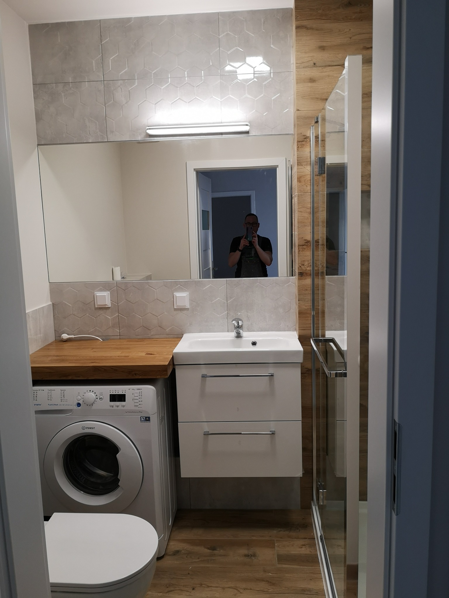 Nowoczesna łazienka po remoncie: szare heksagonalne płytki, drewniana zabudowa pralki, biała umywalka z szafką, kabina prysznicowa ze szklanymi drzwiami, widoczne odbicie fotografa w lustrze.