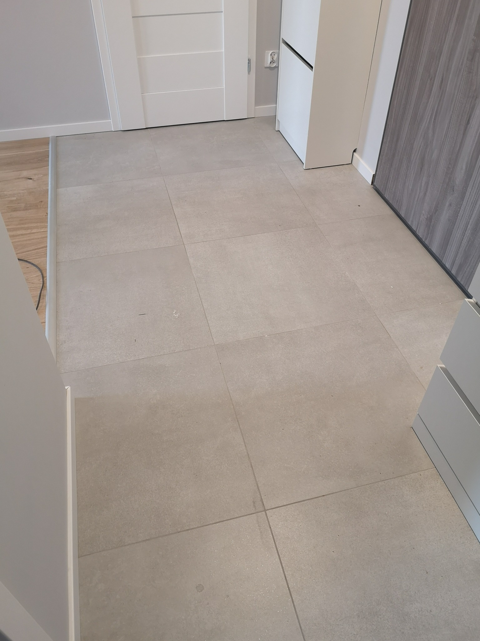 Jasnoszare, matowe płytki podłogowe ułożone w kwadraty, widoczne przy białych drzwiach i fragmentach białych oraz szarych mebli w przedpokoju.