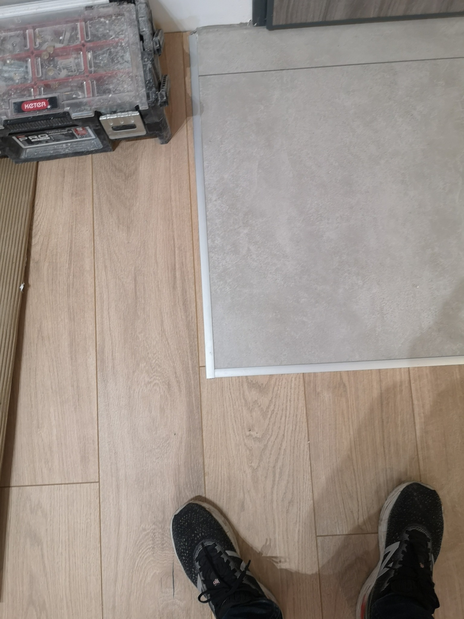 Połączenie paneli podłogowych z płytkami ceramicznymi, widoczna skrzynka narzędziowa Keter i fragment butów sportowych.