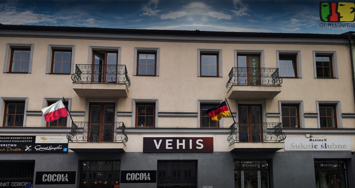 Elewacja kamienicy z balkonami, na których wiszą flagi Polski i Niemiec, nad wejściem szyld 'VEHIS' oraz logo Germanitas w prawym górnym rogu.