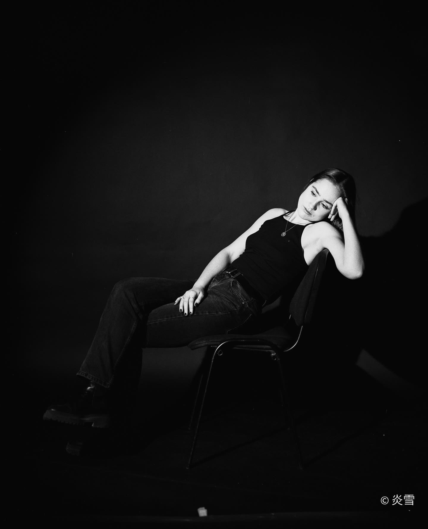 Czarno-białe portretowe ujęcie młodej kobiety siedzącej na krześle na ciemnym tle, zamyślona poza, delikatne światło podkreśla rysy twarzy i fakturę ubrania.