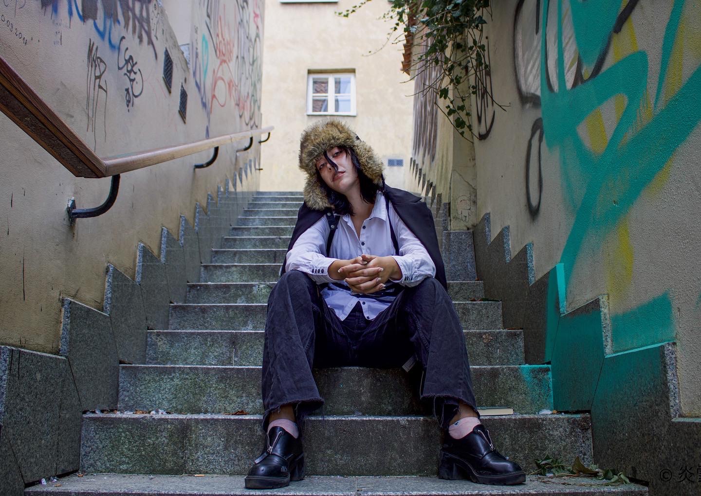 Młoda kobieta w futrzanej czapce i rozpiętej koszuli siedzi na betonowych schodach w wąskim przejściu z graffiti na ścianach, trzymając ręce blisko siebie.