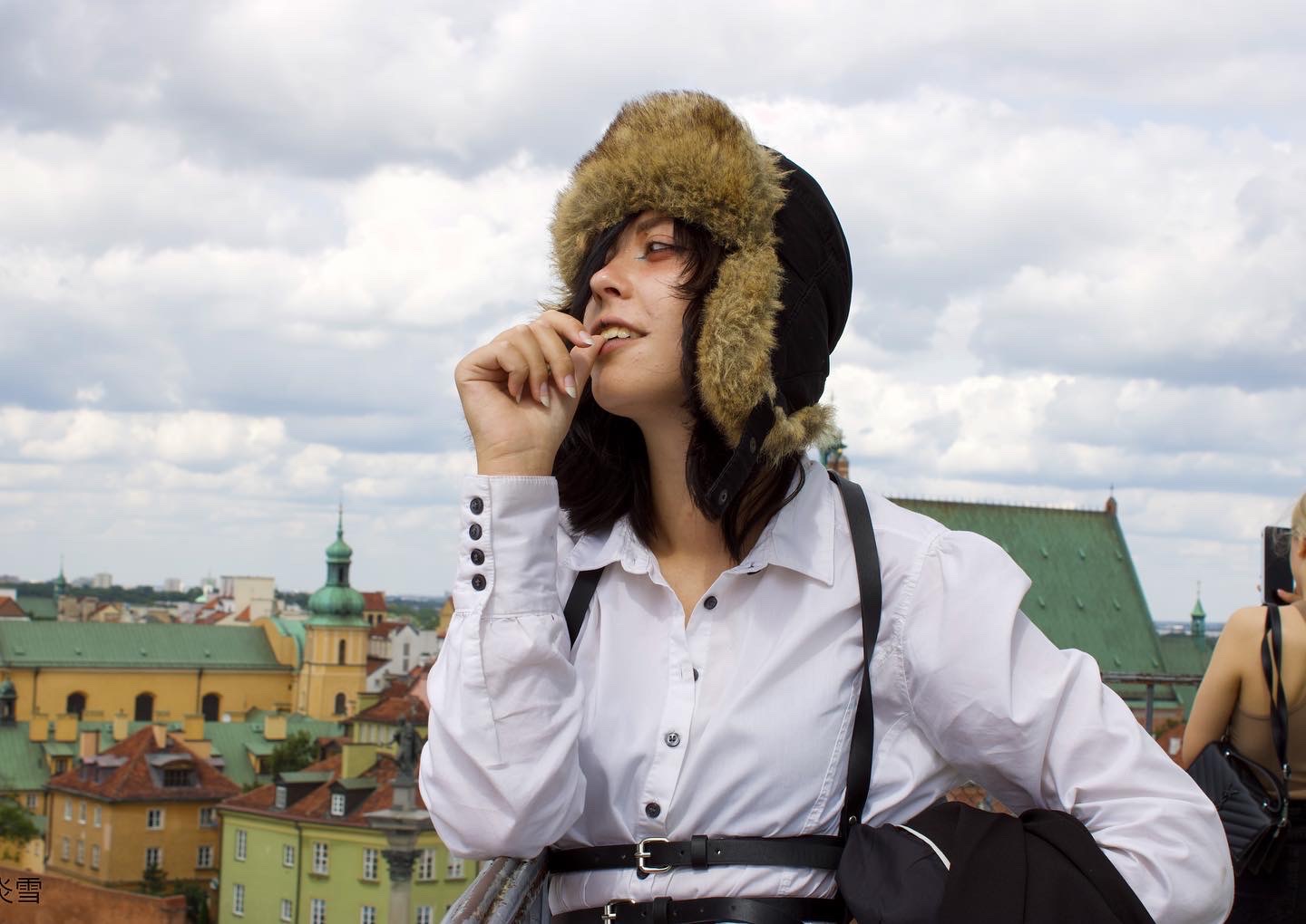 Kobieta w futrzanej czapce i białej koszuli na tle panoramy miasta, z dłonią przy ustach, patrząca w bok.