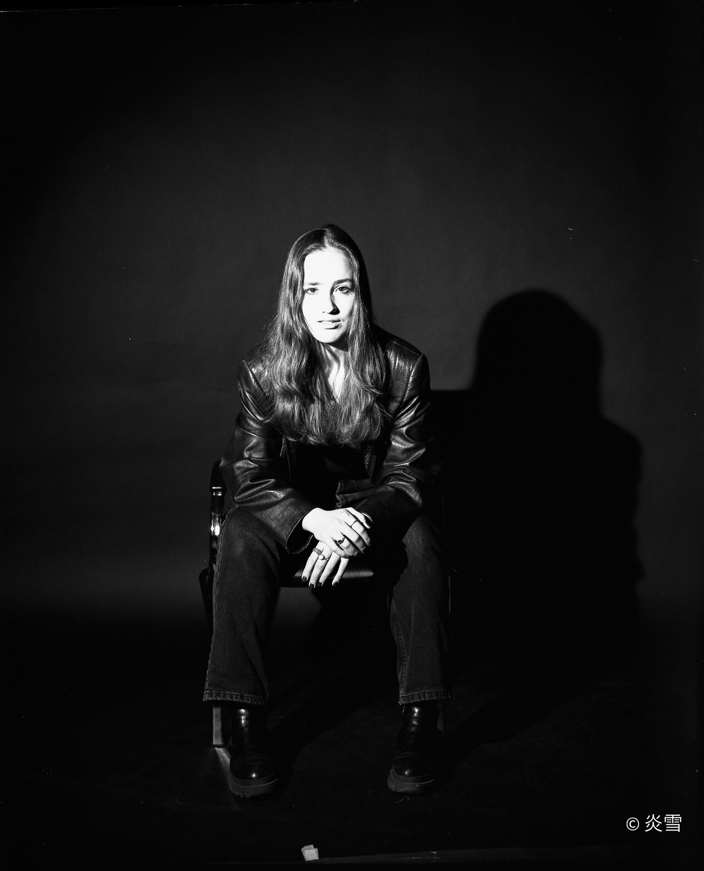 Czarno-białe portretowe ujęcie kobiety siedzącej na krześle w skórzanej kurtce, z dłońmi splecionymi na kolanach, na ciemnym tle z wyraźnym cieniem.