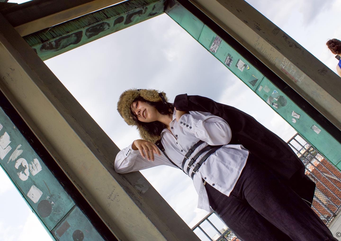 Osoba w futrzanej czapce, białej koszuli z czarnymi paskami i czarnych spodniach, opierająca się o metalową konstrukcję z naklejkami, widziana z dołu pod kątem.