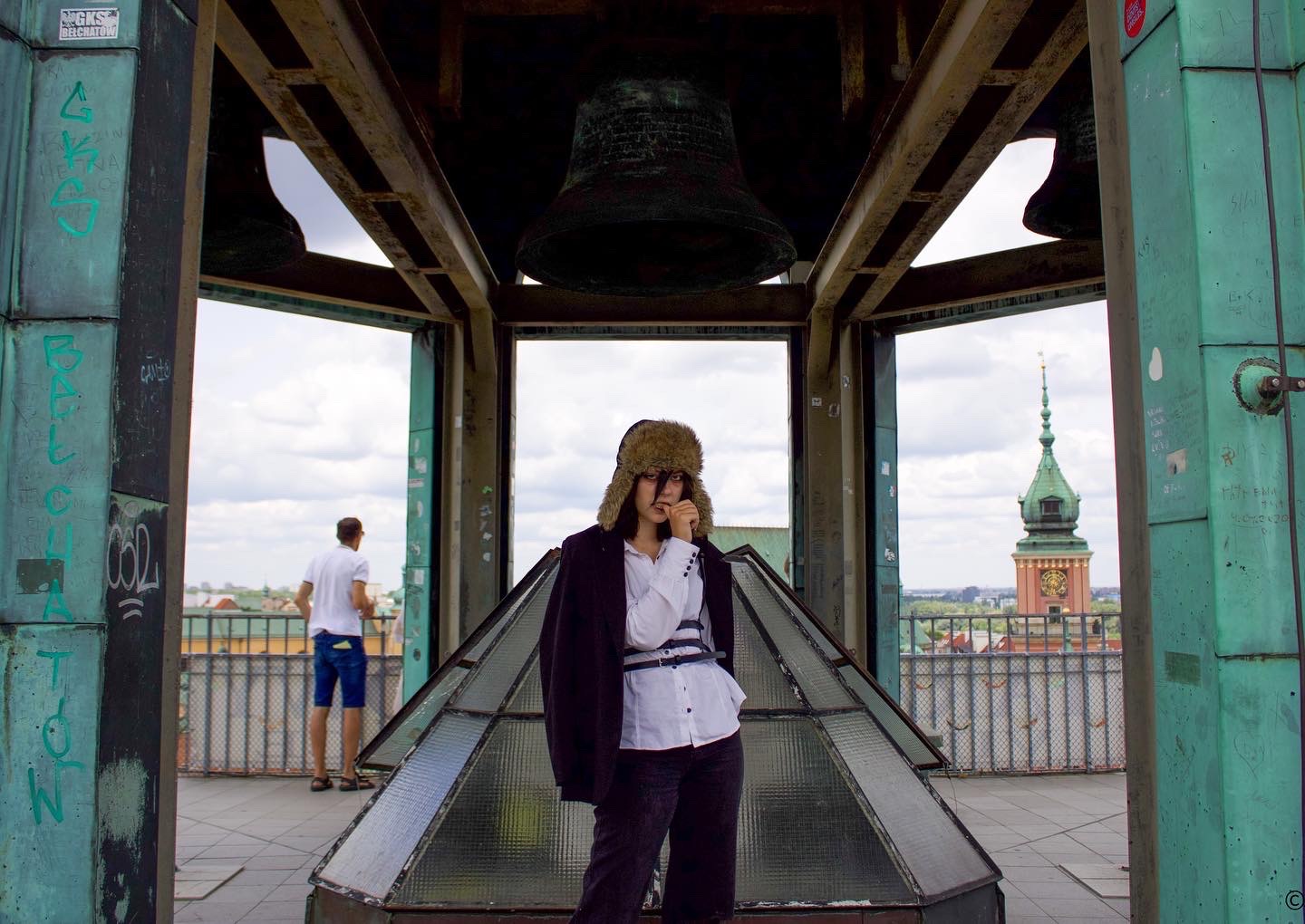 Kobieta w futrzanej czapce i marynarce, stojąca wewnątrz wieży z dzwonami, z widokiem na miasto w tle.