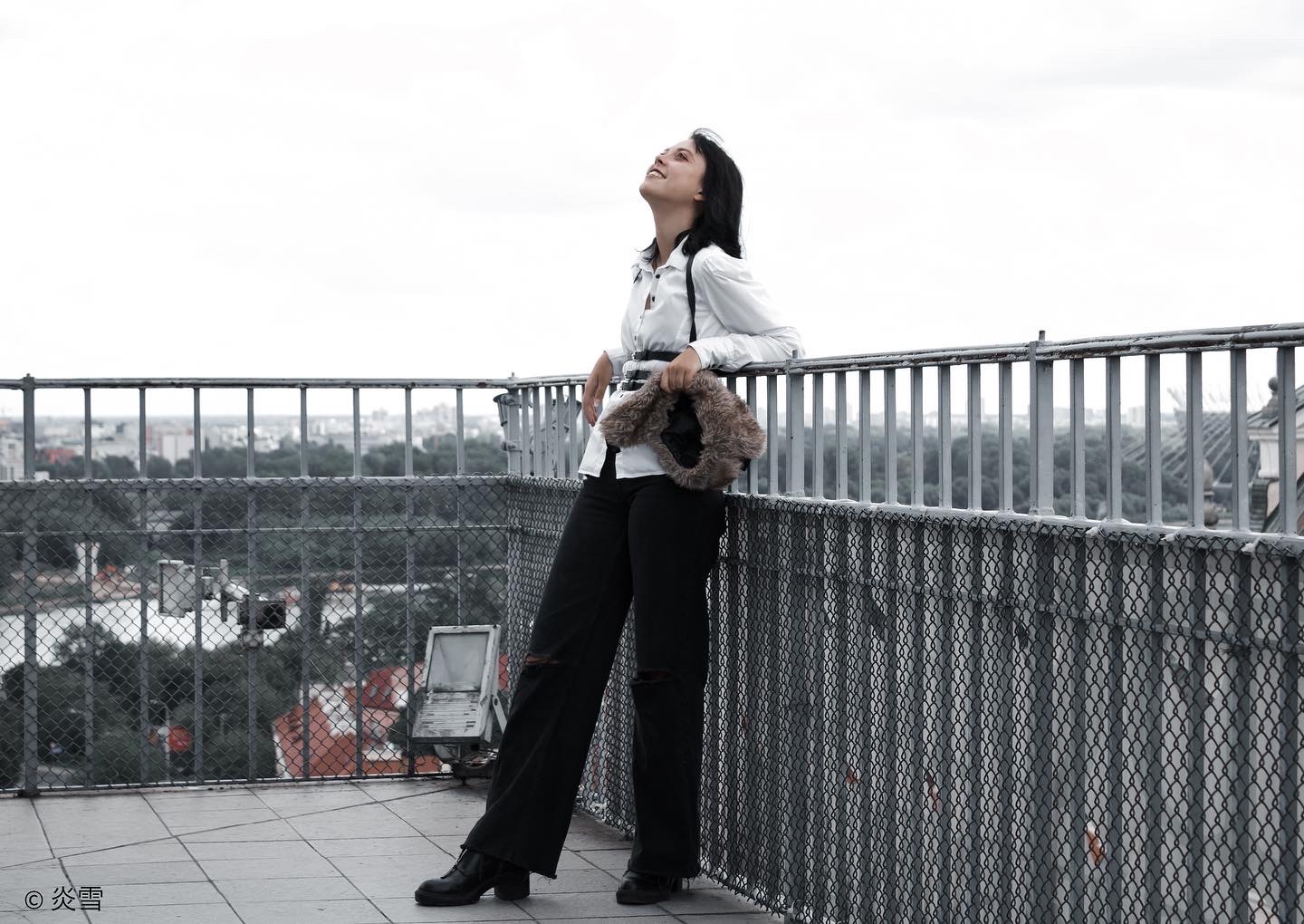 Kobieta w białej koszuli i czarnych spodniach opiera się o metalową barierkę na tle miejskiego krajobrazu, trzymając futrzaną torebkę.