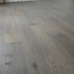 Ułożony parkiet w odcieniach szarości, widoczne fragmenty ściany bez listew przypodłogowych i wystające kable elektryczne.