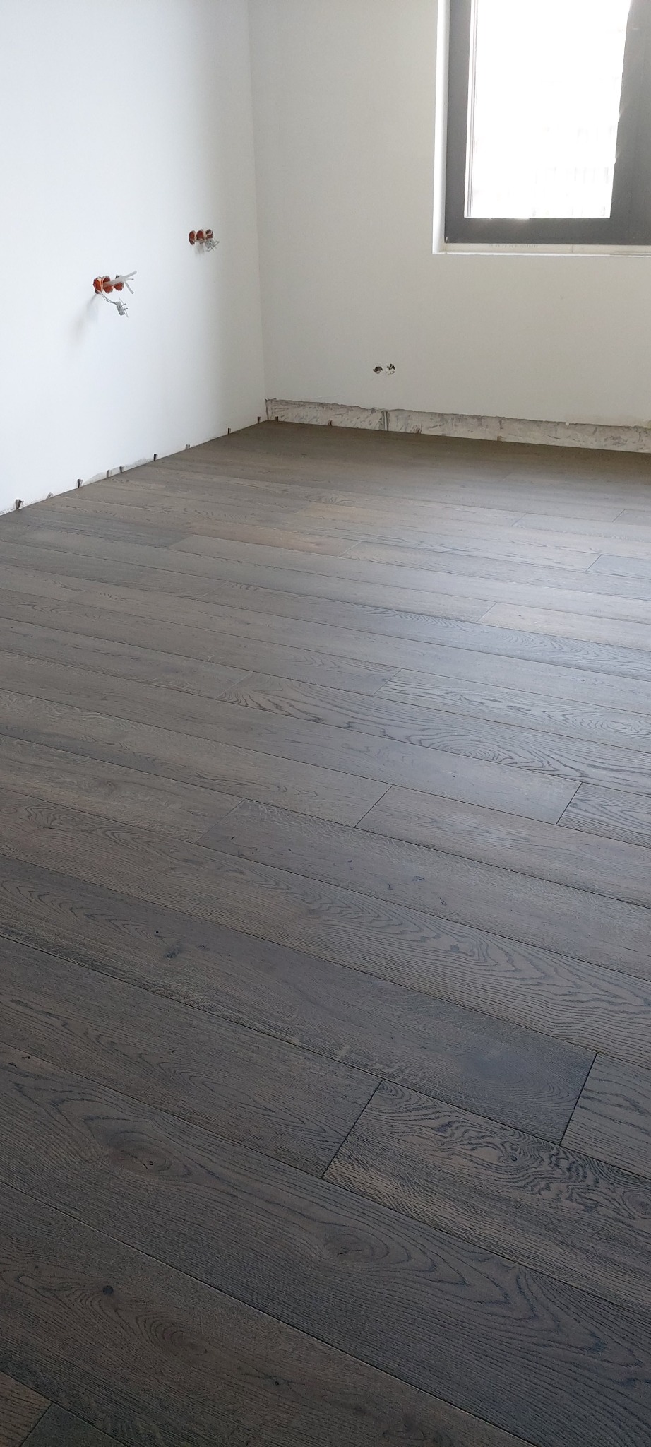 Nowo ułożona, ciemna, dębowa podłoga w pustym pokoju z białymi ścianami i oknem, widoczne wystające kable elektryczne.