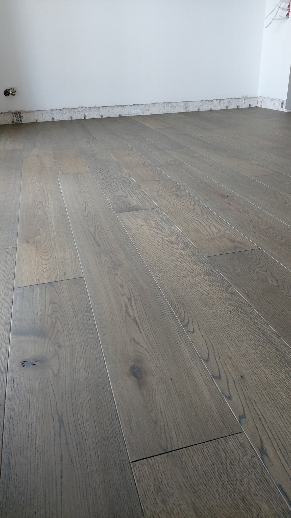 Ułożony parkiet w odcieniach szarości, widoczne fragmenty ściany bez listew przypodłogowych i wystające kable elektryczne.