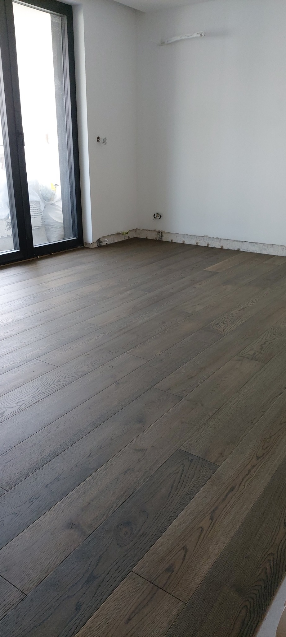 Ułożony parkiet z ciemnego drewna w nowym pomieszczeniu, widoczne niedokończone wykończenia przy ścianach i gniazdka elektryczne.