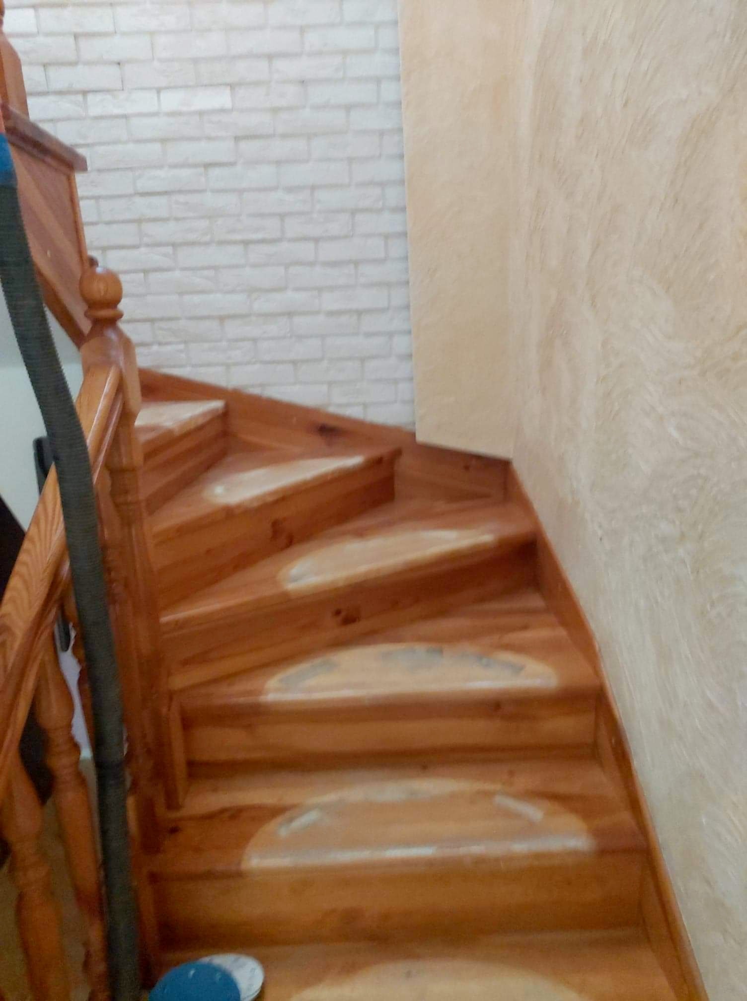 Drewniane schody z balustradą, częściowo wyszlifowane, z widocznym odkurzaczem i ściernicą, na tle ściany z białej cegły i beżowej ściany o fakturze baranka.