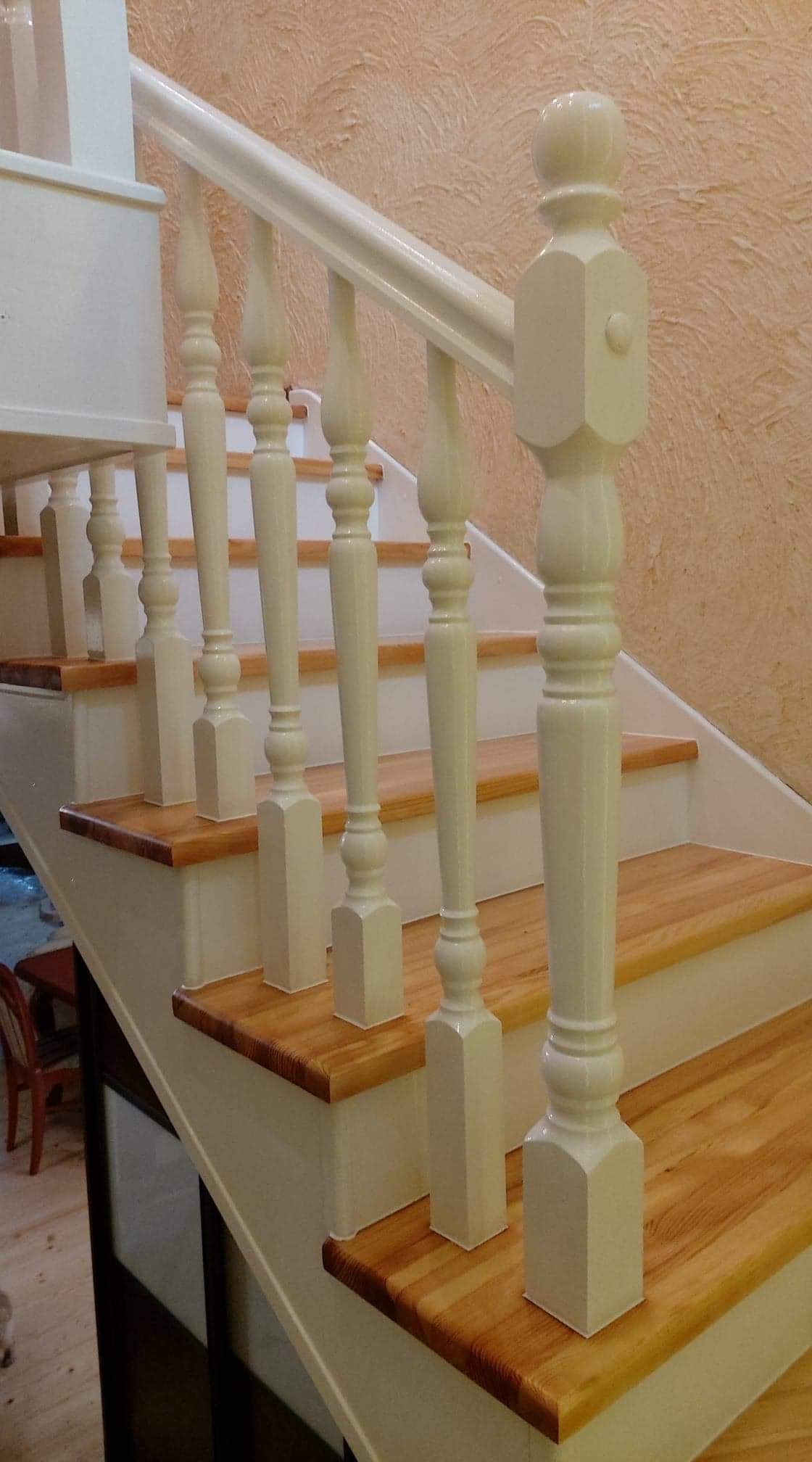 Drewniane schody z białymi balustradami i poręczą, widoczne z dołu, z dekoracyjną fakturą ściany w tle.