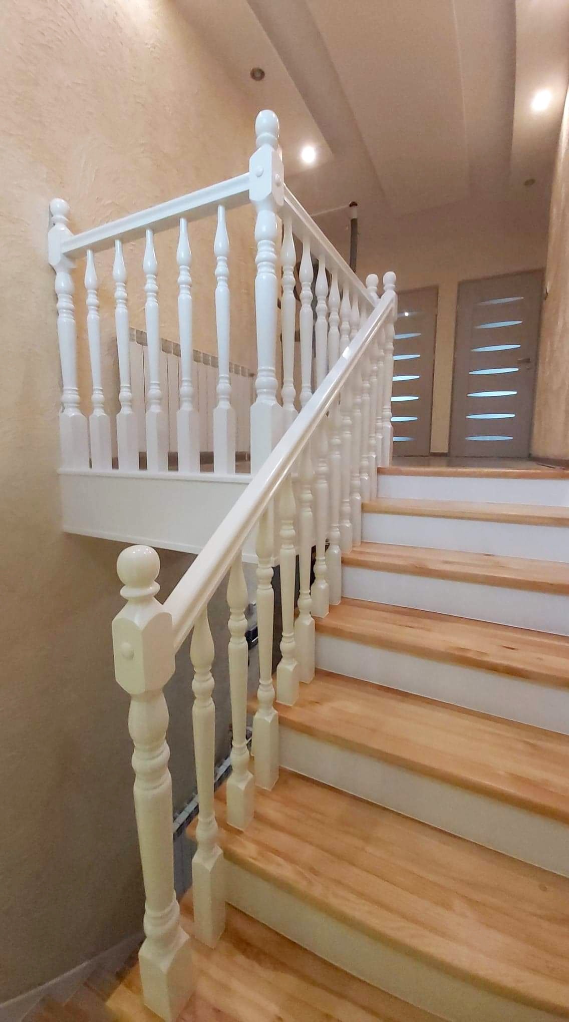 Jasne drewniane schody z białymi podstopnicami i balustradą z toczonymi tralkami, widok z dołu na górę, drzwi w tle.