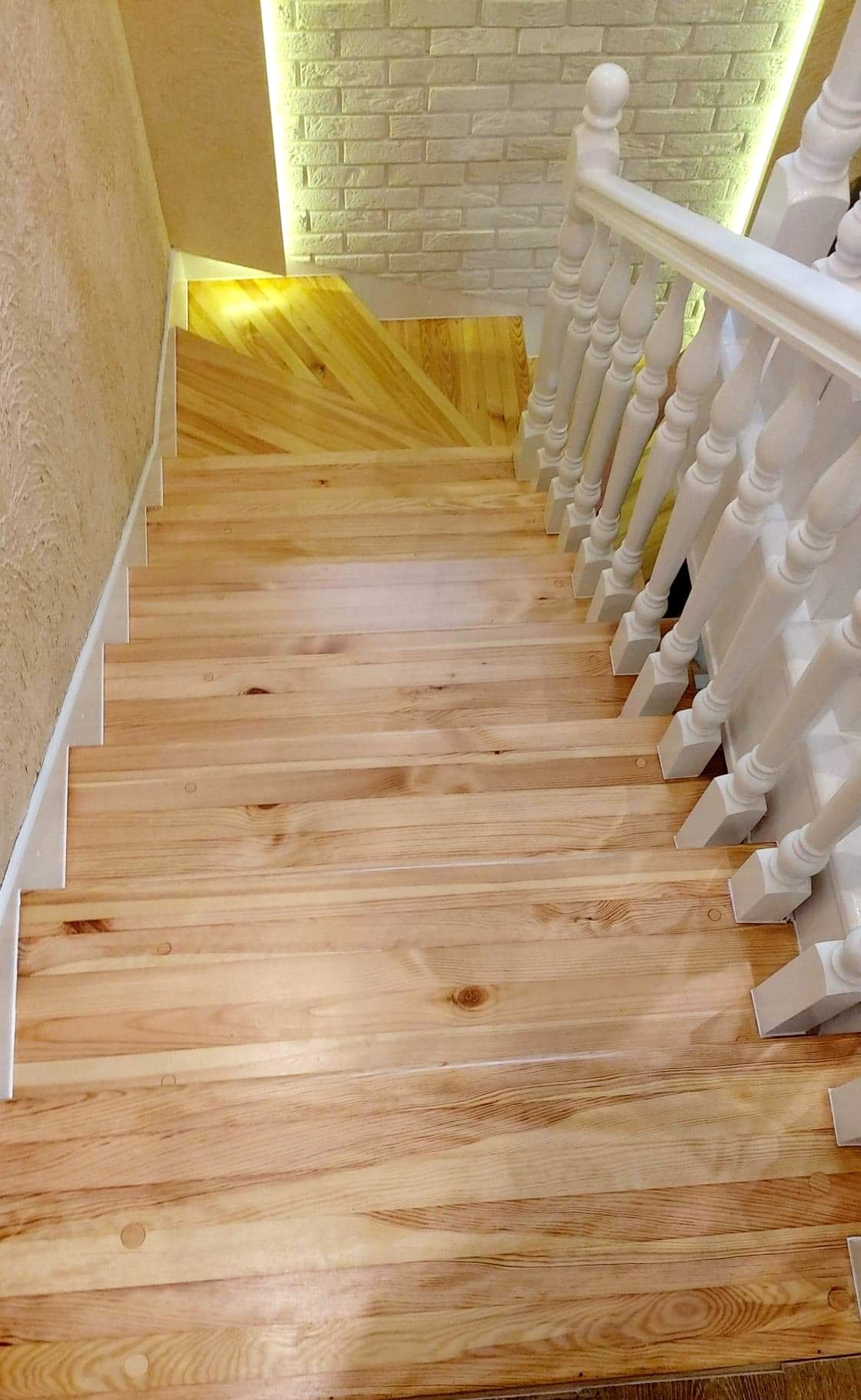 Widok z góry na drewniane schody z białymi balustradami, wykończone jasnym drewnem, z fragmentem ściany z cegły dekoracyjnej w tle.