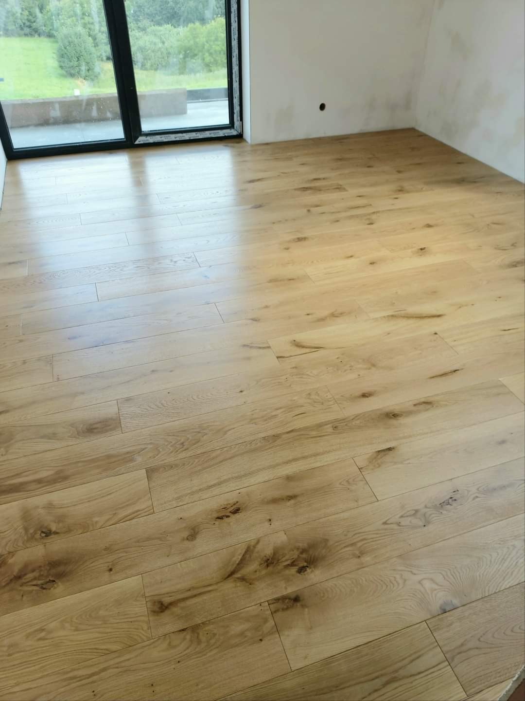 Jasne, drewniane panele podłogowe ułożone w nowym pokoju z dużym oknem balkonowym, ściany przygotowane do malowania.