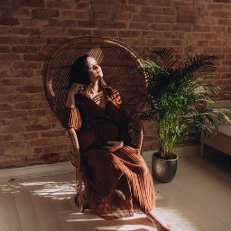 Nietuzinkowa fotografia Katarzyna Wesołowska - Kobieta w brązowej sukience ciążowej siedzi w wiklinowym fotelu na tle ceglanej ściany, oświetlona promieniami słońca, obok doniczkowa roślina.