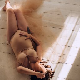 Nietuzinkowa fotografia Katarzyna Wesołowska - Kobieta w cielistym body leży na białej drewnianej podłodze z delikatną, beżową tkaniną, z ręką na brzuchu i tatuażem na ramieniu, oświetlona naturalnym światłem.