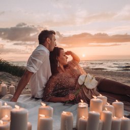 Nietuzinkowa fotografia Katarzyna Wesołowska - Para leży na plaży przy zachodzie słońca, otoczona zapalonymi świecami i kwiatami anturium, kobieta w błyszczącej sukience patrzy w dal.