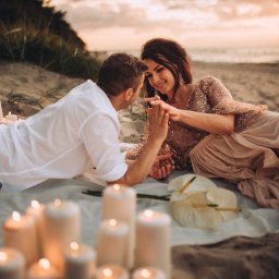Nietuzinkowa fotografia Katarzyna Wesołowska - Para zakochanych leży na kocu na plaży o zachodzie słońca, otoczona świecami i białymi kwiatami, trzymając się za ręce.