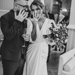 Nietuzinkowa fotografia Katarzyna Wesołowska - Para młoda w czerni i bieli, pokazująca obrączki, ona w sukni z dekoltem V i tatuażami, on w okularach i garniturze, oboje trzymają kieliszki z szampanem.