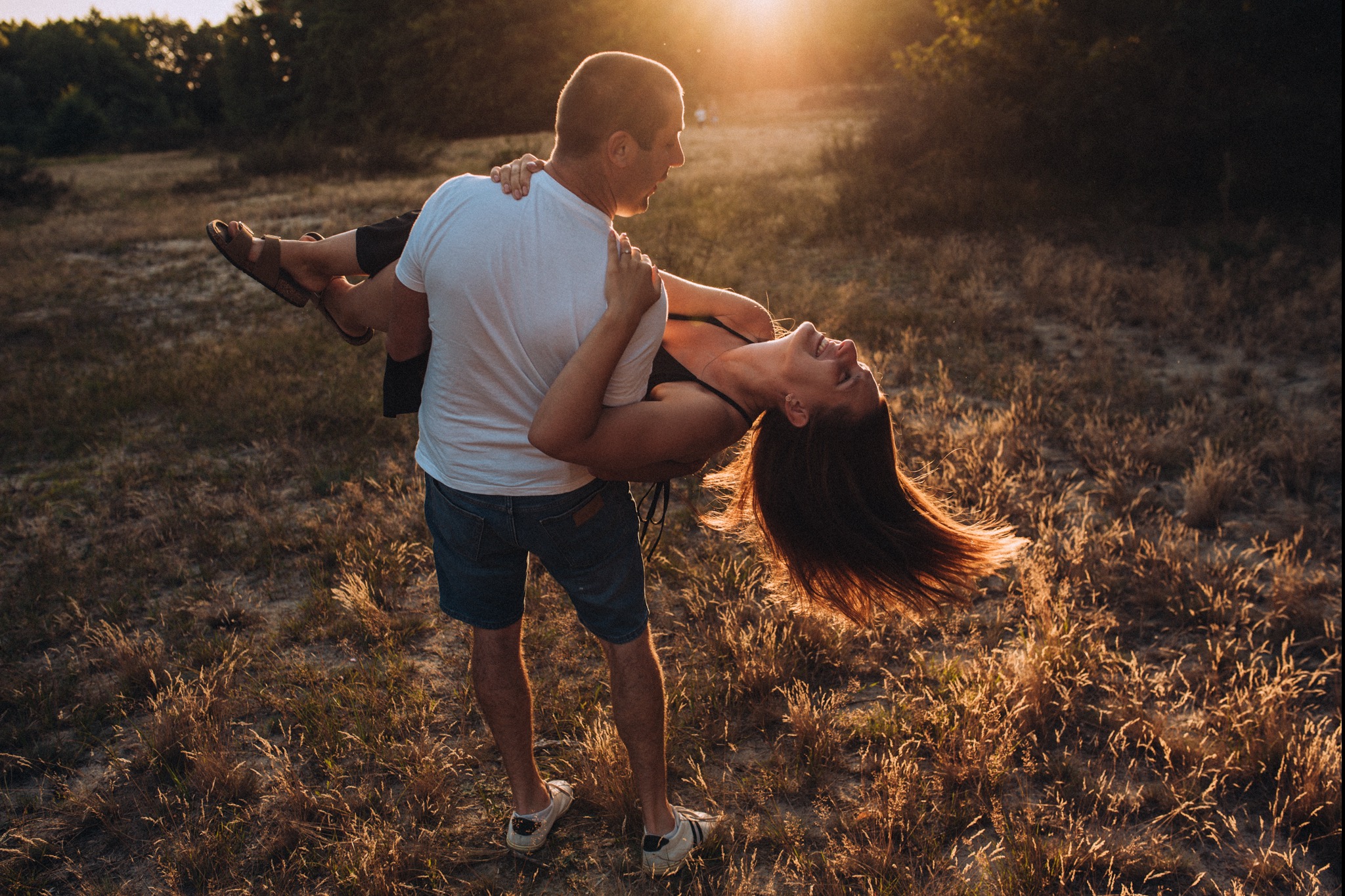 Mężczyzna trzyma kobietę w ramionach na tle zachodzącego słońca, oboje uśmiechnięci, w naturalnym otoczeniu łąki.