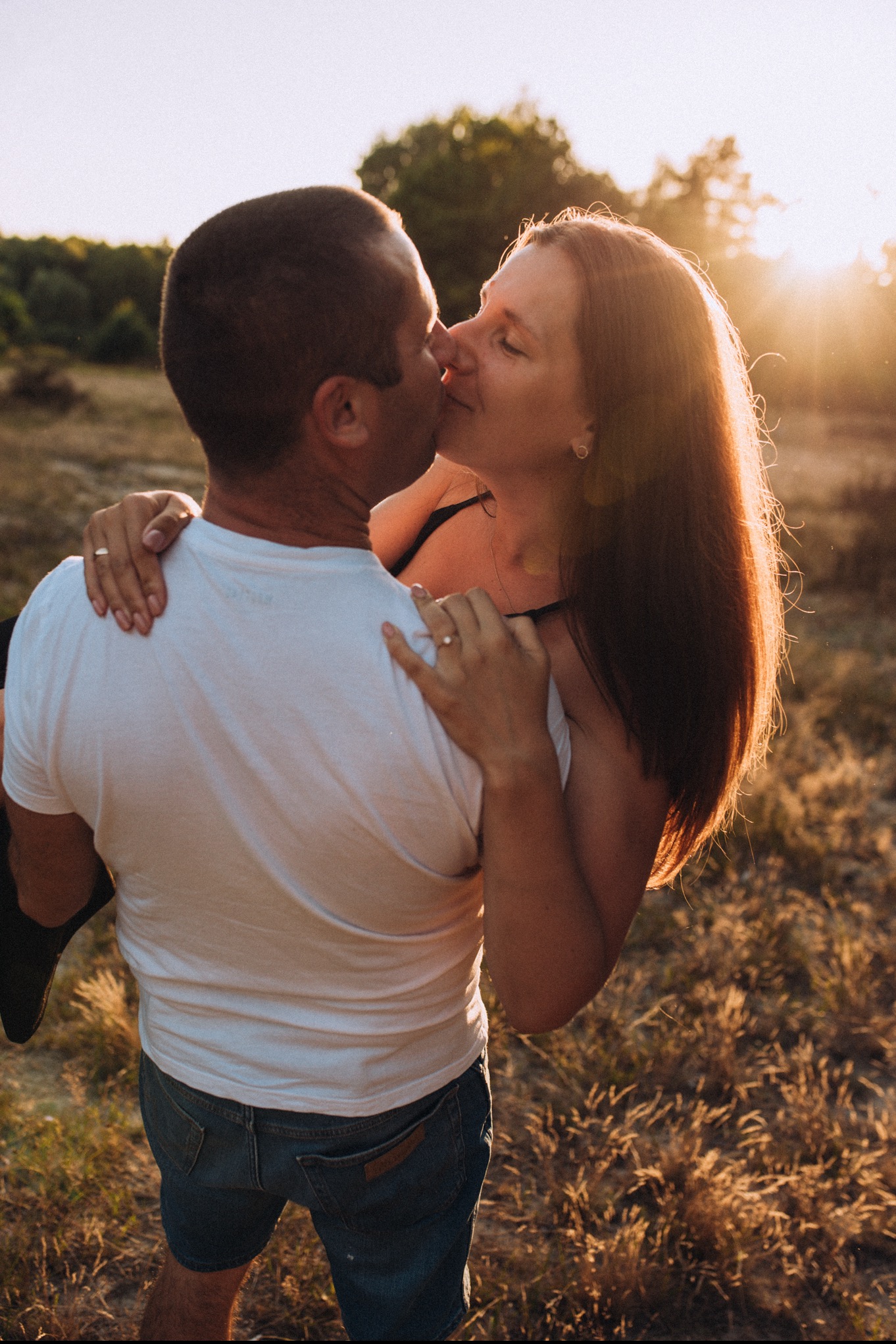 Zakochana para całuje się na tle zachodzącego słońca, mężczyzna trzyma kobietę w ramionach, oboje mają na palcach obrączki