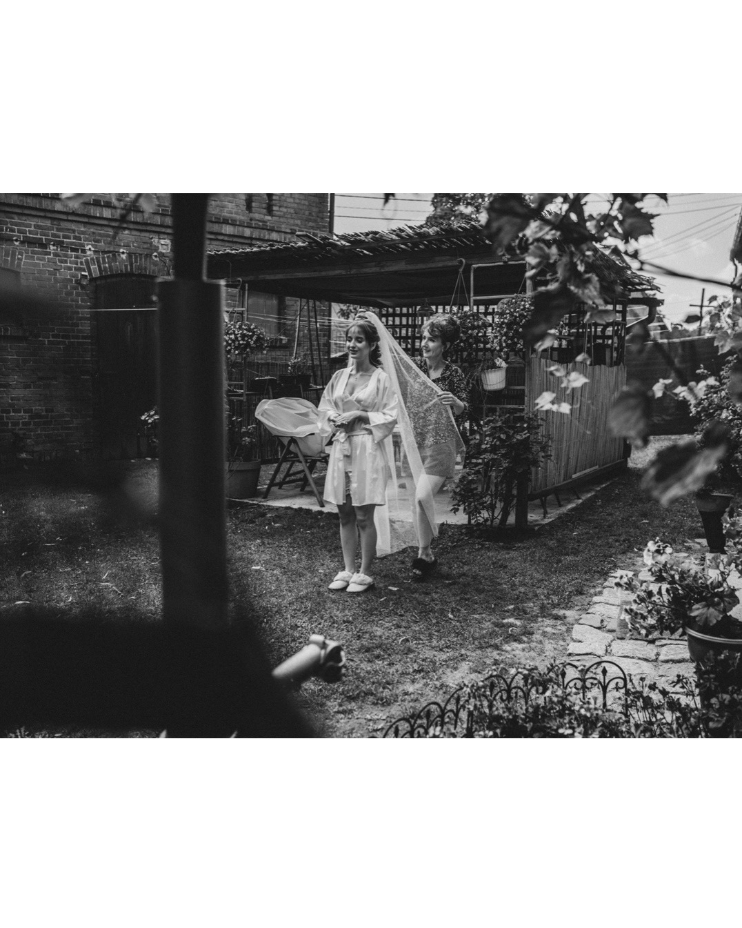 Czarno-białe zdjęcie panny młodej w szlafroku i welonie, poprawianym przez drugą kobietę w ogrodzie przed domem z cegły.