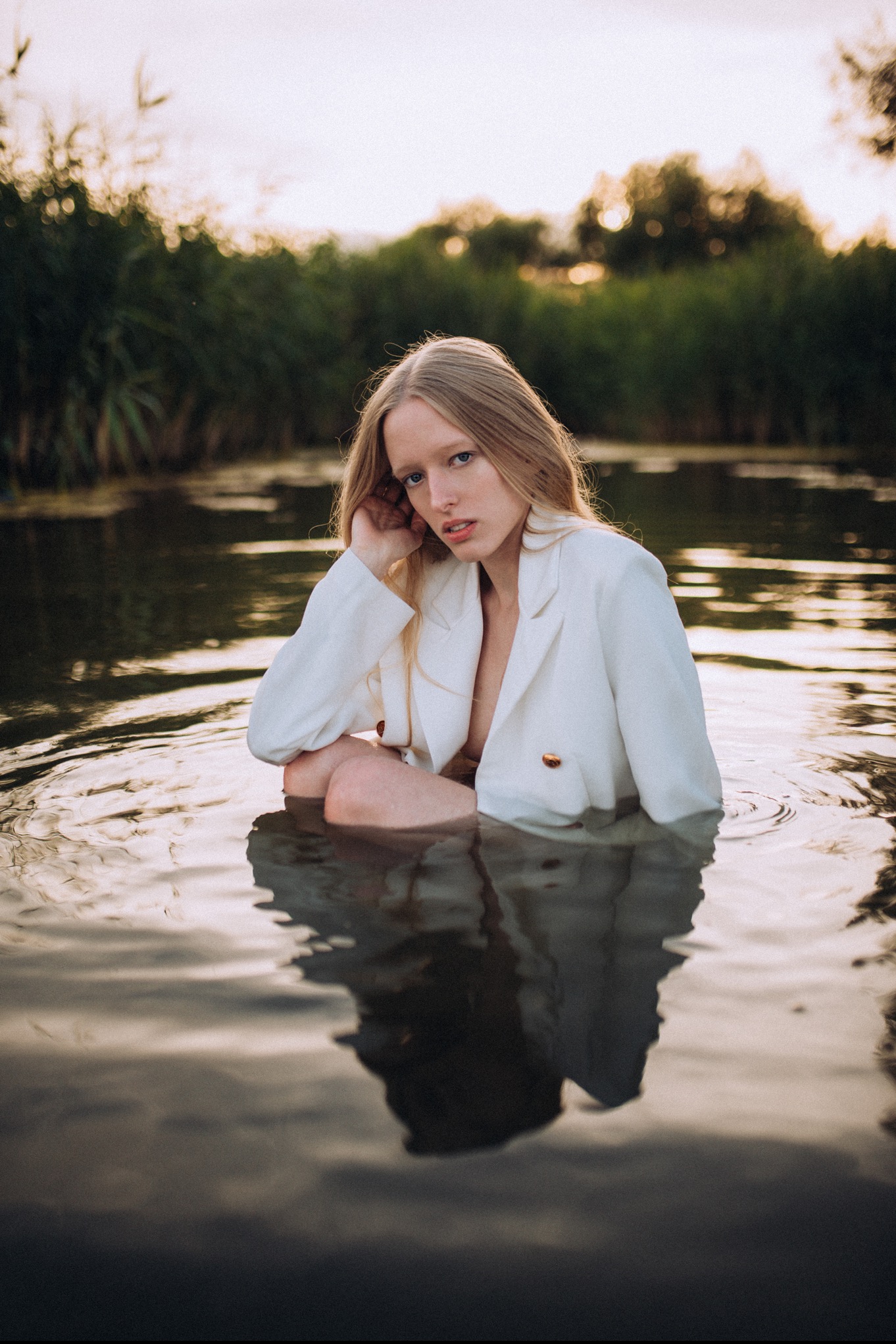 Kobieta w białej marynarce zanurzona w wodzie, z trzcinami i drzewami w tle, delikatne oświetlenie wieczorne.