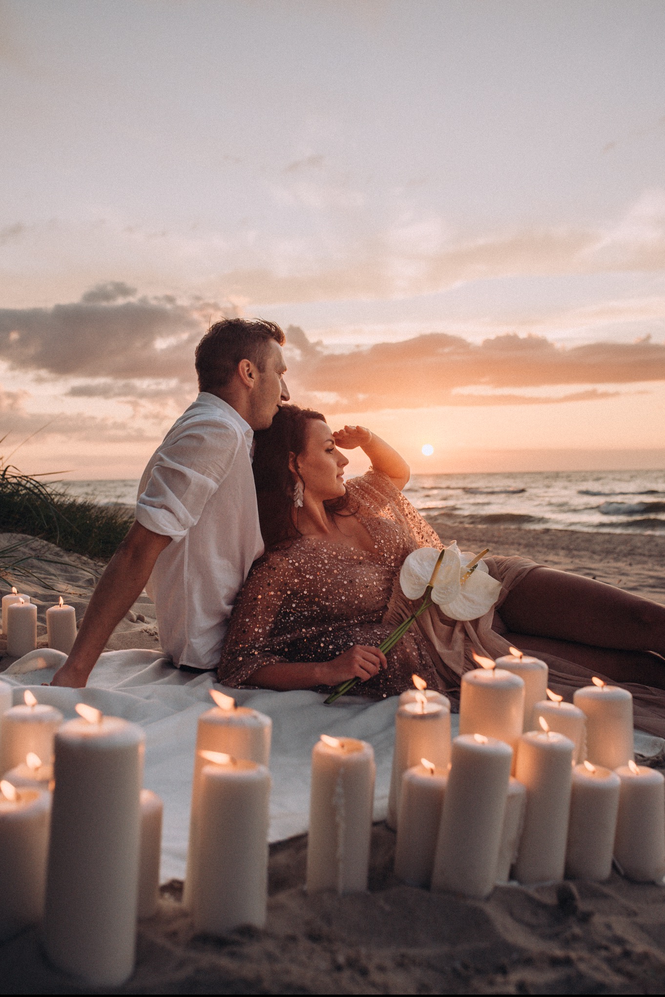 Para leży na plaży przy zachodzie słońca, otoczona zapalonymi świecami i kwiatami anturium, kobieta w błyszczącej sukience patrzy w dal.