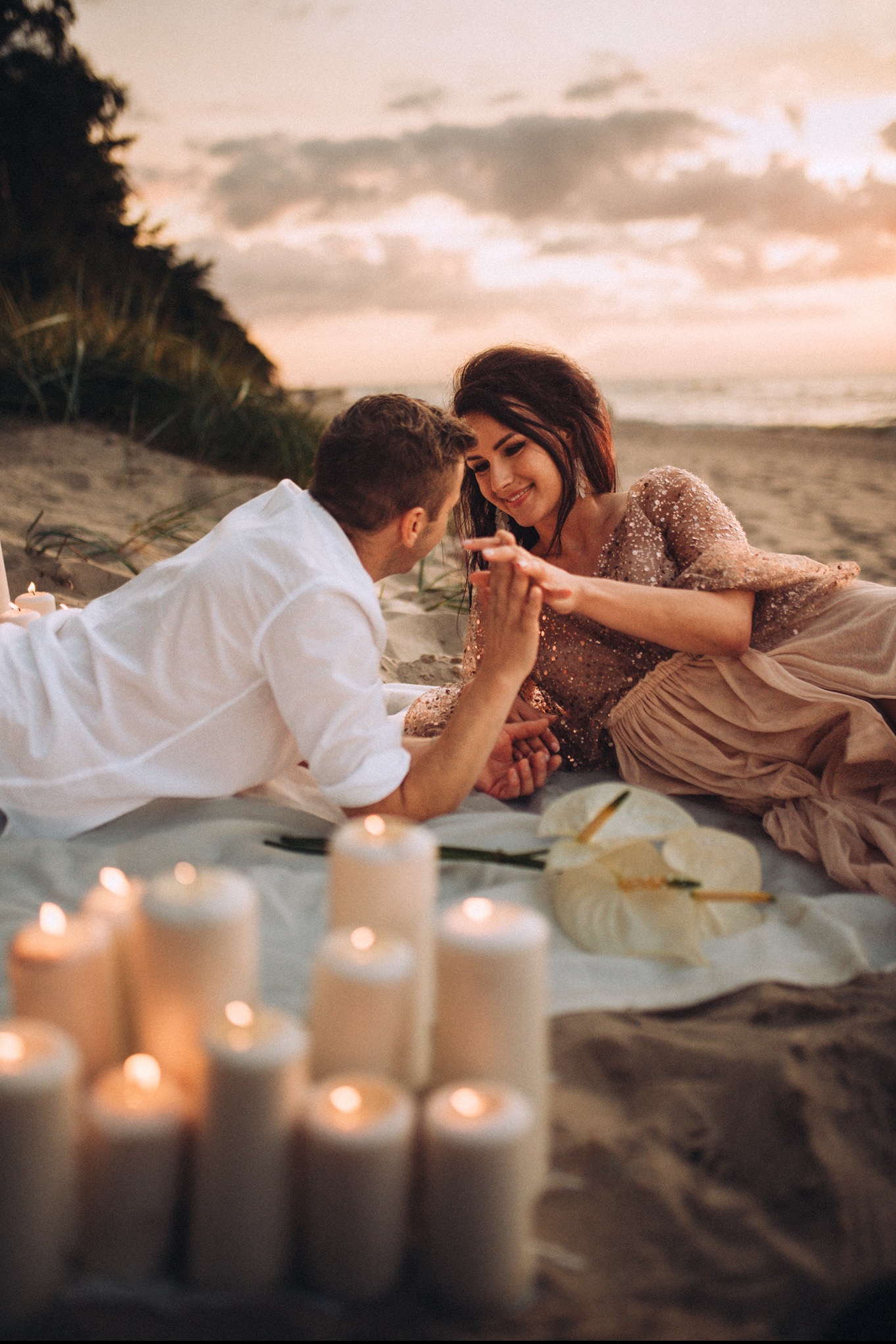 Para zakochanych leży na kocu na plaży o zachodzie słońca, otoczona świecami i białymi kwiatami, trzymając się za ręce.