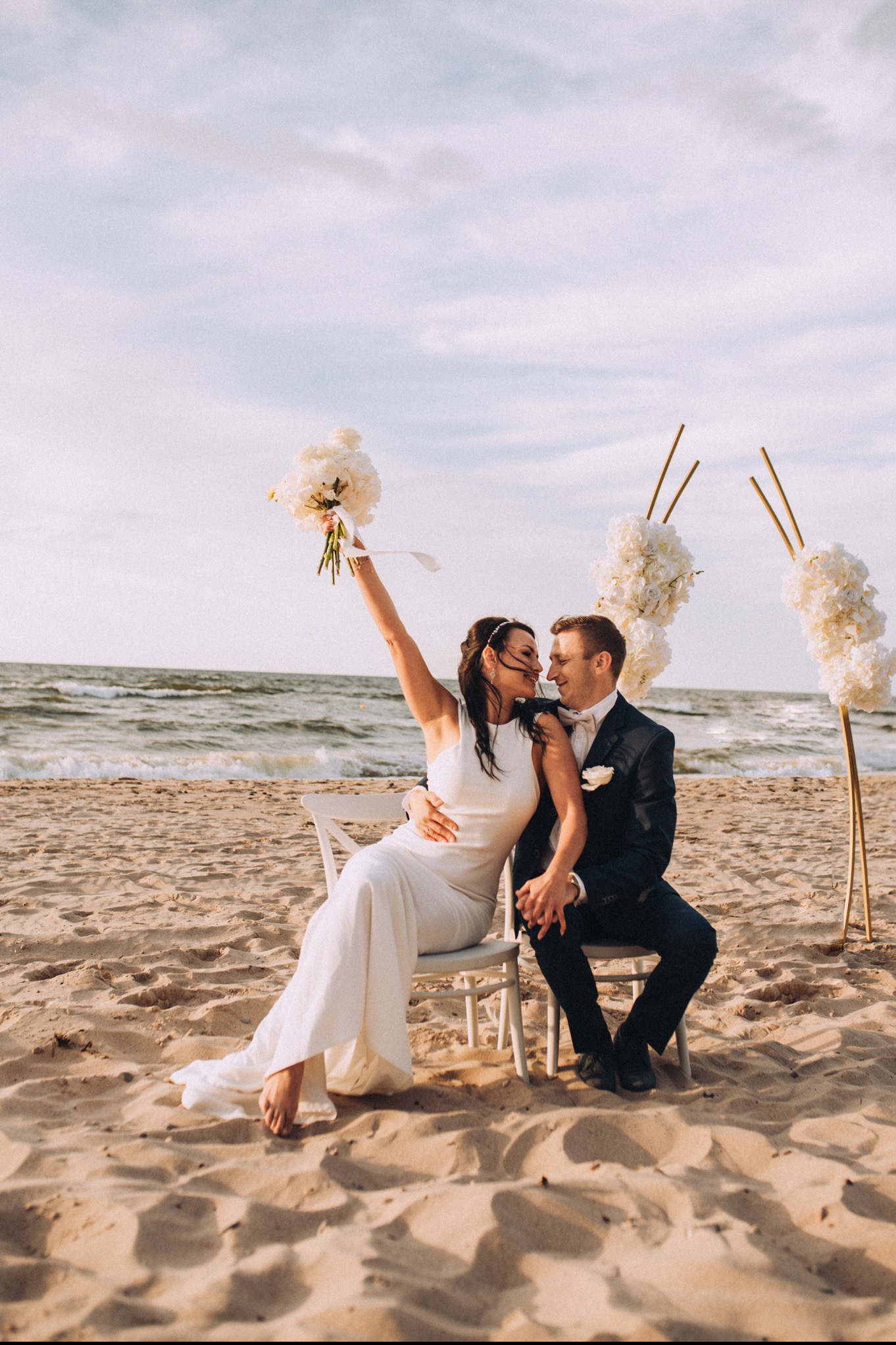 Para młoda siedzi na białych krzesłach na plaży, panna młoda trzyma bukiet białych kwiatów, w tle morze i dekoracje kwiatowe na złotych stelażach.