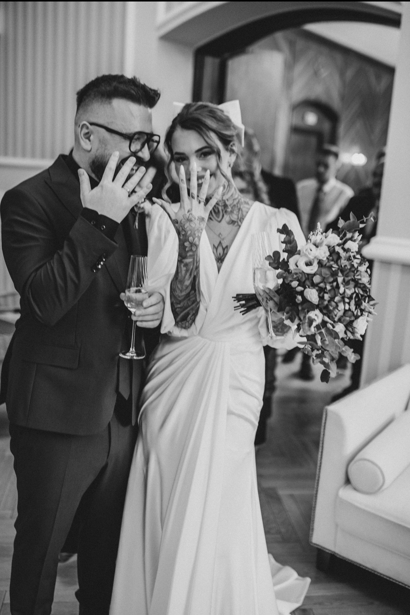 Para młoda w czerni i bieli, pokazująca obrączki, ona w sukni z dekoltem V i tatuażami, on w okularach i garniturze, oboje trzymają kieliszki z szampanem.